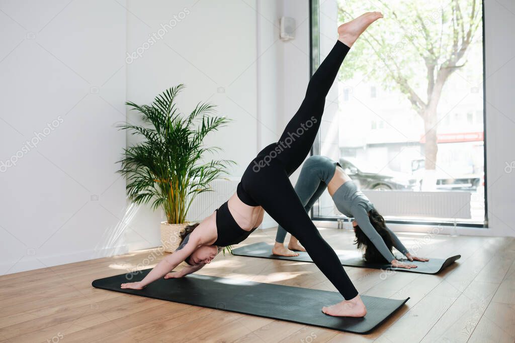 Focused women in sportswear practicing yoga in a group, doing downward facing dog in a big studio. Side view