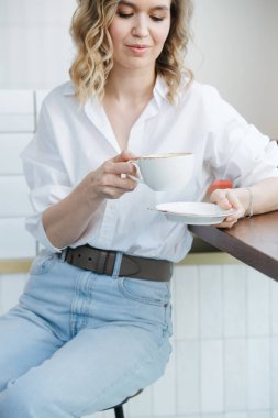 Kafede yüksek bir taburede oturan yalnız sarışın kadın bir fincan kahveye bakıyor. Üzerinde mavi gömlek ve açık renk kot pantolon var..