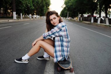 Boş bir şehir yolunun ortasında kaykayın üzerinde oturan düşünceli bir kadın. Kareli gömlek ve mini kot pantolon giyiyor..