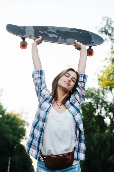 Tired happy woman stretching with a skateboard in hands. Low angle. She's wearing open checkered shirt atop regular white one. Eyes closed.