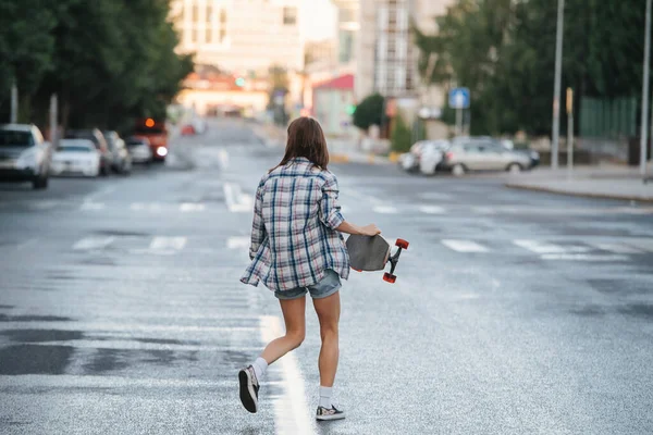 女性が早朝に手にスケートボードでぬれた空の都市の道路を歩いています チェック柄のシャツとミニジーンズのショートパンツを着ている 顔を背けて — ストック写真