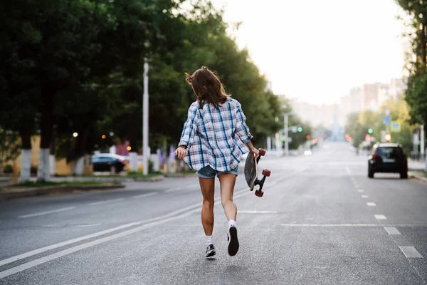 早朝に手にスケートボードでぬれた空の都市の道路を歩いて不思議な女性 チェック柄のシャツとミニジーンズのショートパンツを着ている 顔を背けて — ストック写真