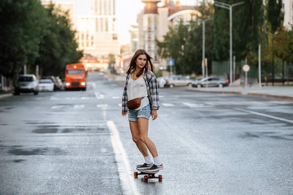 早朝に空の街の道路上のスケートボードに乗ってかなり若い女性 カメラを見てください チェックのシャツとミニジーンズのショートパンツを着てる — ストック写真