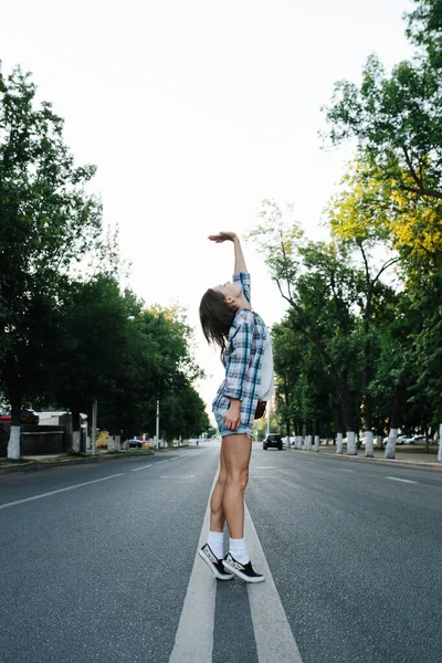 奇妙な若い女性が空の街の道路の真ん中にストレッチ 低い角度だ チェッカーの開いたシャツを着てる サイドビュー — ストック写真