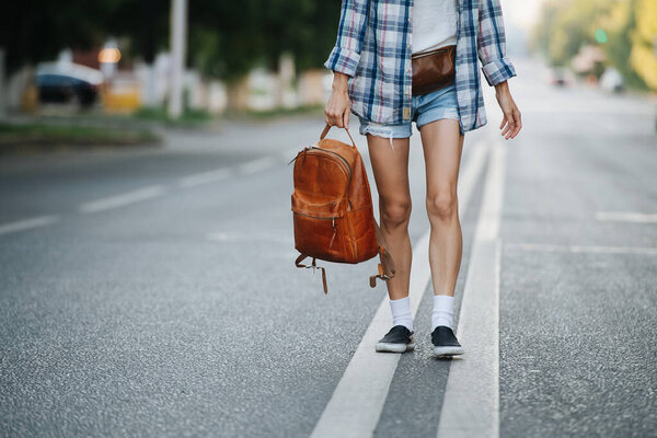 Lower part of a woman walking on the road, holding brown leather backpack. No head. She's wearing checkered shirt and mini jeans shorts.