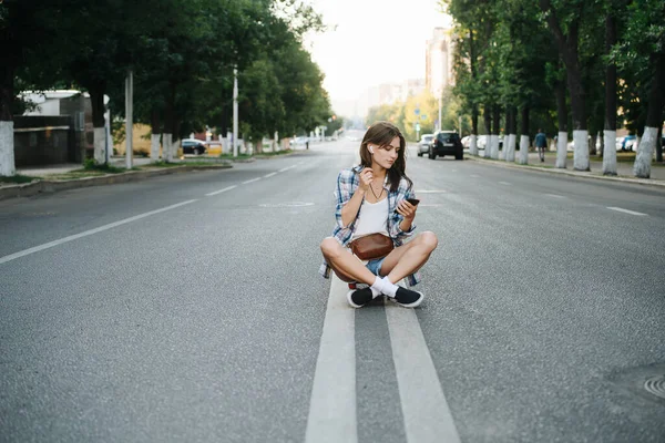 平和な無責任な女性は 空の街の道路の真ん中に 手に彼女の携帯電話で座っている 音楽を聴く チェックのシャツとミニジーンズのショートパンツを着てる — ストック写真