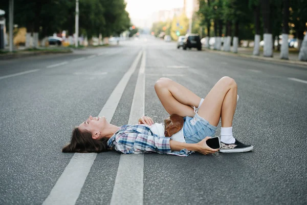 狂気の光アスファルトの道路に横たわって若い女性は 状況について笑っている サイドビュー チェックのシャツとミニショートパンツを着てる — ストック写真