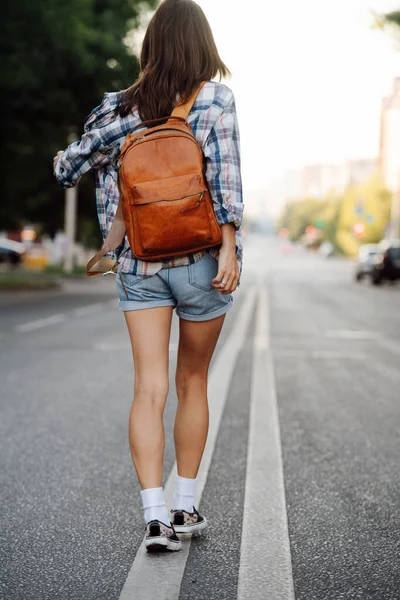 ダブル分割線に沿ってアスファルトの道路を歩くブルネットの女性は 茶色の革のバックパックを着て チェックのシャツとミニジーンズのショートパンツを着てる — ストック写真