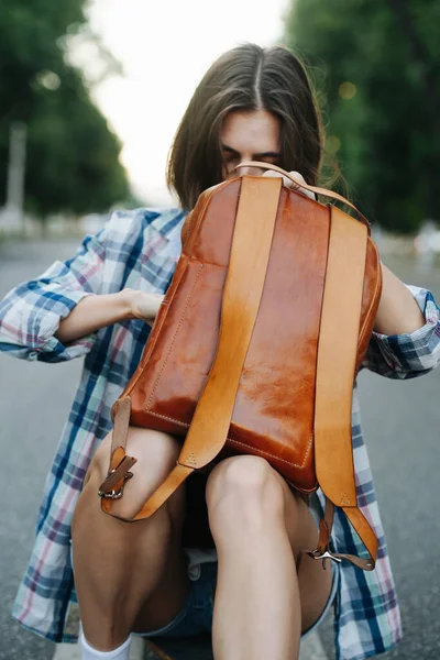 女性はアスファルトの上に座って 彼女の茶色の革のバックパックの中に何かを探している チェックのシャツとミニショートパンツを着てる — ストック写真