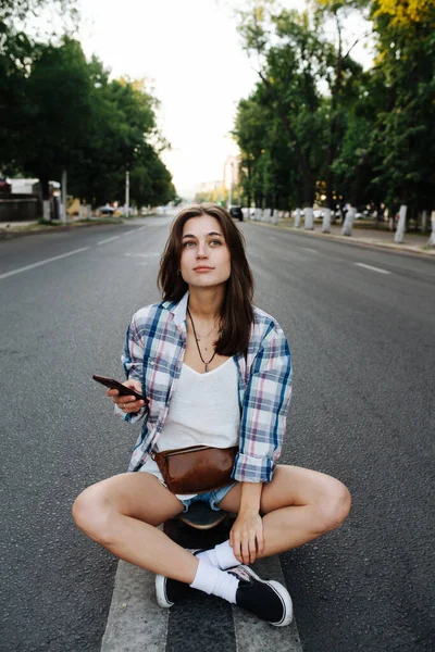 良い女性は 空の都市道路の真ん中に 手に彼女の携帯電話で座っている チェックのシャツとミニジーンズのショートパンツとレザーのファニーパックを着てる — ストック写真