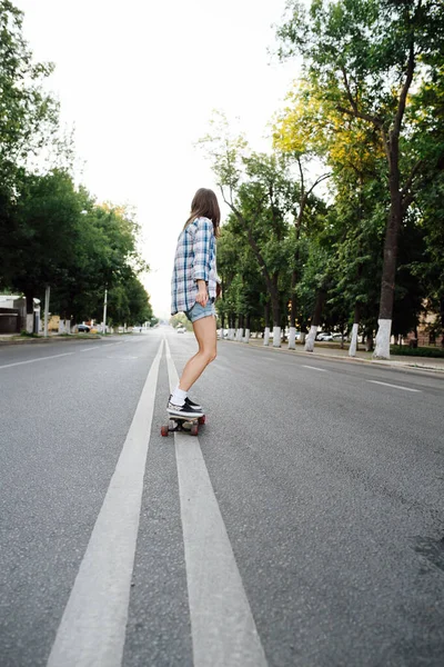 一位年轻女子坐在空荡荡的城市道路上的滑板上 与一个很好的平衡骑着走的低角度图像 她穿着格子衬衫和迷你裙短裤 脸转过去 — 图库照片