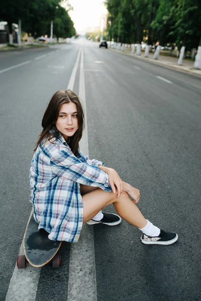 きれいな女性は 空の街の道路の真ん中にスケートボードに座っている 気をつけて彼女の考えに満足してる チェックのシャツとミニジーンズのショートパンツを着てる — ストック写真