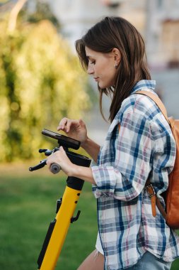 Parkın kontrol ayarlarındaki esmer kadın elektrikli scooter kiraladı. Yan görüş. Kareli gömlek ve mini kot pantolon giyiyor..
