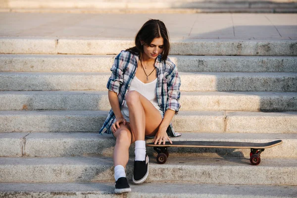 幸せな女性は階段に座って 愛とケアで彼女のスケートボードを見下ろしています 穏やかな日差しの下で チェックのシャツとミニジーンズのショートパンツを着てる — ストック写真