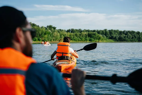 People in orange life jackets kayaking on a big wide river. There are several boats close and afar. Second seat view.