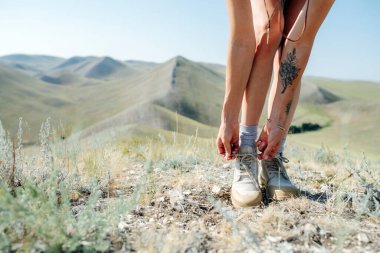 Bir kadın resmi, öne eğiliyor, ayakkabılarını bağlıyor. Arkasındaki tepe manzarası çok güzel. Kesildi, kafası yok..