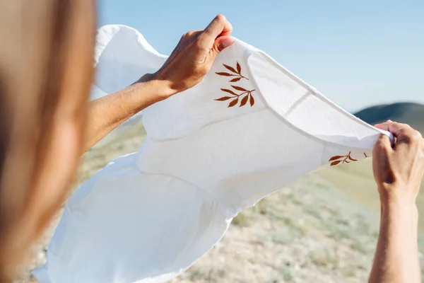 Woman in a countryside holding in hands a white dress, fluttering in the wind. Hands and hair only. From behind.