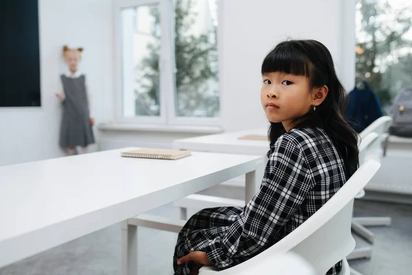一个身穿格子制服的亚裔女学生坐在课桌后面的画像 看着摄像机Look Camera 模糊的背景 — 图库照片