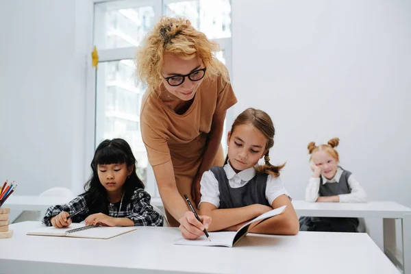 Profesora Ayudando Una Colegiala Sentada Detrás Escritorio Escribiendo Cuaderno Vista — Foto de Stock