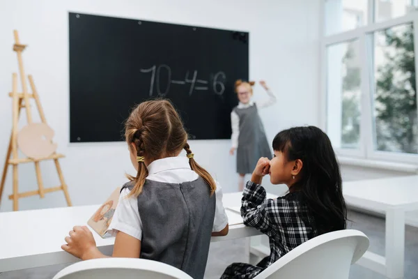 两个小女孩坐在书桌后面 看着一本华丽的书 他们的同学在模糊的背景下在黑板上解数学等式 — 图库照片