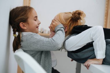 Aktif ilkokul kızları birbirleriyle oynuyorlar. Biri yüze girmek, ikincisi avuçlarla gözlerini kapamak. Yan görünüm.