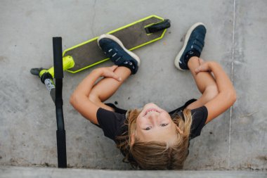 Beton zeminde motosikletin yanında oturan gülümseyen bir çocuğun en üst görüntüsü. Kameraya bakıyorum..