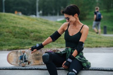 Güvertede dinlenen kısa saçlı, gülümseyen bir kadın patencinin düşük açılı görüntüsü. Koruyucu giysi giyiyor. Ön manzara. Arkaplanda yeşil park.