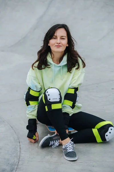 コンクリートの床の上に彼女のボードに座っているスポーツウェアのブルネットスケーター 彼女はカメラを見てる — ストック写真