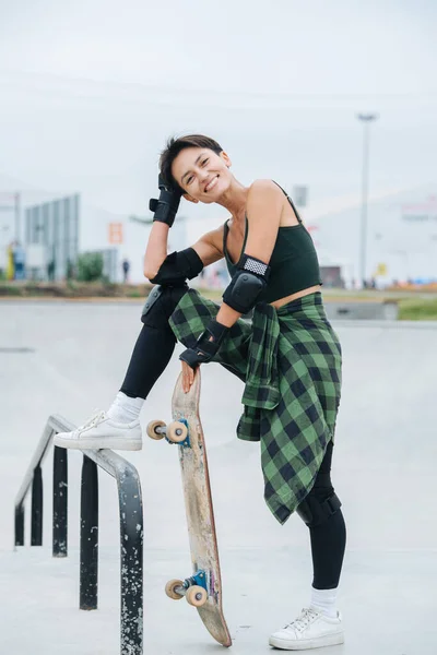 明るいスケーターの女性が手すりに1本足で歩き ボードにもたれている姿が描かれている 黒んぼの街を背景に — ストック写真