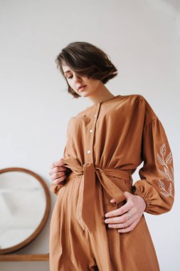 Kahverengi tulumlu, kemerini sıkan seksi bir kadının yukarı açılı görüntüsü. Güzel esmer, içeride kısa saçlı, yuvarlak aynalı bir masanın yanında.