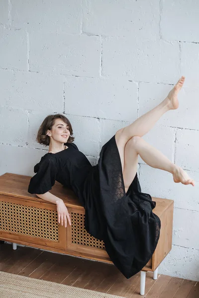 Flirty playful young woman lying on a low cupboard in a modest black dress. Assuming provocative pose, lifting her bare legs up. Sexy brunette with short hair indoors.