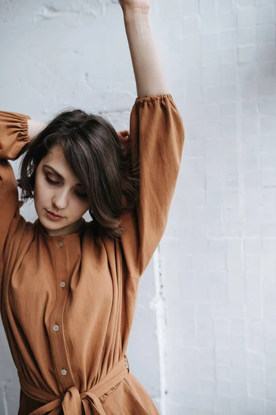 Sensual woman in a brown jumpsuit stretching, lifting arms, looking down. Beautiful brunette with short hair indoors.