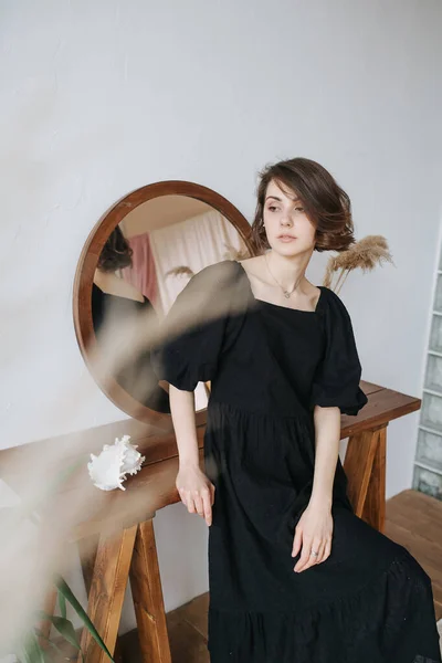 Pretty languid woman in a black dress leaning on a table with a round mirror. Beautiful brunette with short hair indoors.