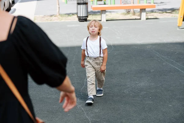 Crying four year old boy walking on asphalt to his mother for hugs and comfort. From behind woman\'s, shoulder.