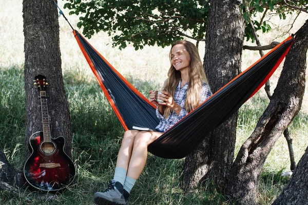 Mujer alegre sentada en una hamaca, entre dos grandes árboles. Beber té — Foto de Stock