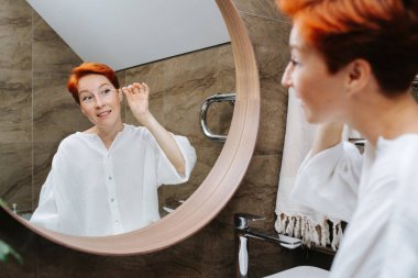 Kızıl saçlı olgun kadın göz makyajını pamukla düzeltiyor. Banyoda aynanın karşısında dikiliyor..