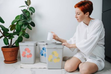 Odaklanmış kızıl saçlı kadın bacaklarının üzerinde oturuyor, çöpleri evdeki küçük geri dönüşüm kutularıyla ayırıyor. Üzerlerinde farklı renkte oklar var. Plastik bardak tutarak.