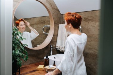 Güzel, kısa saçlı, olgun bir kadın saçlarını ahşap bir tarakla tarıyor. Banyoda aynanın önünde dikiliyor, odak noktasında yansıması var..