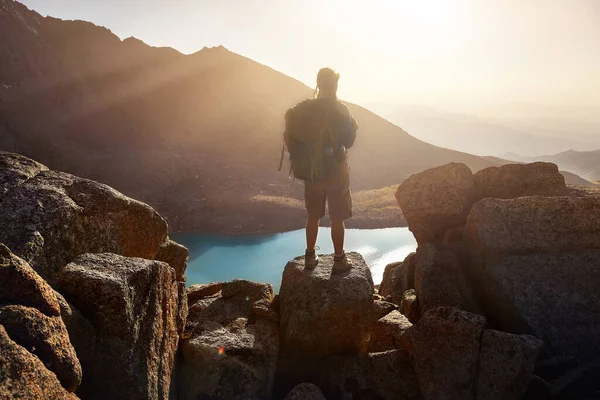 在山谷的湖畔 背着背包站在岩石上 美丽的落日映衬着人类远足游客的轮廓 户外及远足的概念 — 图库照片