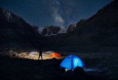 Erkek turist gece gökyüzünün altındaki dağlarda kamp yaparken yıldızlarla eldiven çadırını aydınlatıyor.