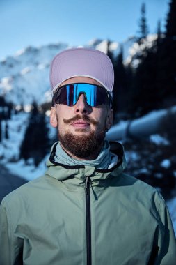 Kışın güneşli bir günde dağ yolunda mor şapkalı ve güneş gözlüklü sakallı bir sporcunun portresi. Spor giysileriyle doğada koşmak..