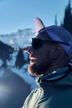 Kışın güneşli bir günde dağ yolunda mor şapkalı ve güneş gözlüklü sakallı bir sporcunun portresi. Spor giysileriyle doğada koşmak..