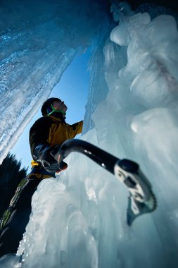 Sarı ceketli adam büyük şelalede buz baltası ve kramponlarla donmuş buz duvarına tırmanıyor. Kış dağlarında mavi kayağa karşı. Spor dağcılığı ve dağcılık konsepti