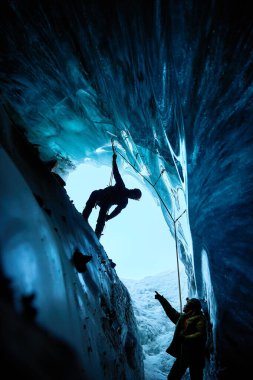 Siyah silüetli birkaç dağcı, buz baltası ve kramponlarla buz dağlarının içindeki donmuş buz mağarasına tırmanıyor. Spor dağcılığı ve dağcılık konsepti