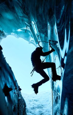 Siyah siluetli adam buz baltası ve kramponlarla buz dağlarındaki donmuş buz mağarasına tırmanıyor. Spor dağcılığı ve dağcılık konsepti