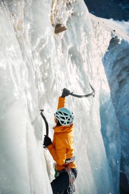 Turuncu ceketli, mavi kasklı ve baltalı genç bir Asyalı atlet. Buz tırmanış takımları, Kazakistan 'ın Almaty kenti yakınlarındaki dağlarda dikey donmuş şelaleye tırmanıyor.