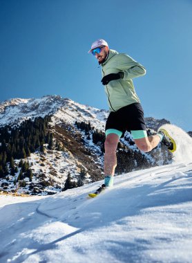 Erkek sporcu kış güneşli bir günde dağ vadisinde kar yolunda. Natur 'da spor kıyafetleriyle koşuyor.