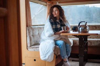 Karavanda beyaz bir battaniyeyle bir masada oturan ve elinde kupa tutan bir kız.