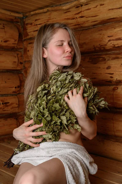 Jeune femme relaxante dans un sauna vêtue d'une serviette et d'un balai de bouleau — Photo