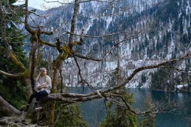 Kız ağaçta oturur ve mavi göl arka planında meditasyon yapar. Abhazya 'daki Ritsa Gölü